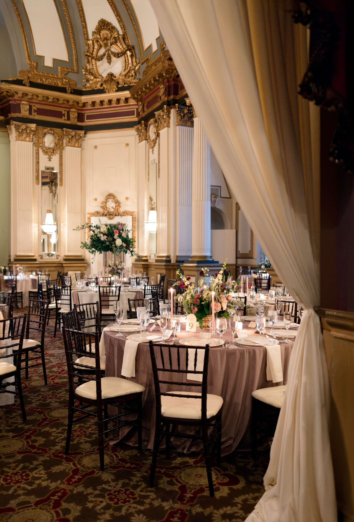 A lavish ballroom set for an event, featuring elegantly decorated tables with floral centerpieces and candles. The room is adorned with ornate architectural details, high ceilings, and soft lighting, creating an ambiance of sophistication and elegance.