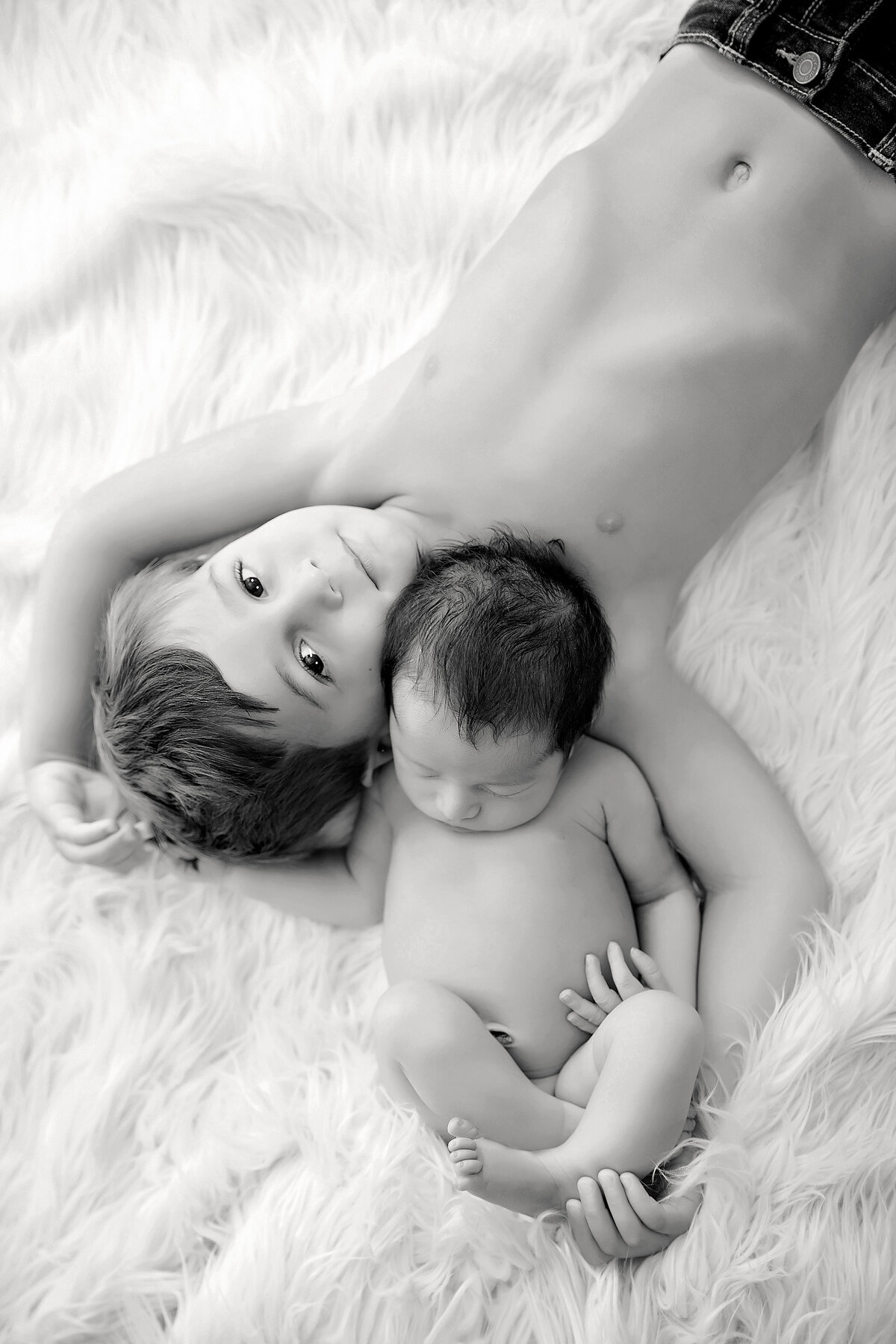 Black and white image by a Jacksonville baby photographer captures a young child and a baby lying on a fluffy white rug. The older child, with short hair and wearing shorts, cradles the baby in their arms. Both have serene expressions, gazing upwards.