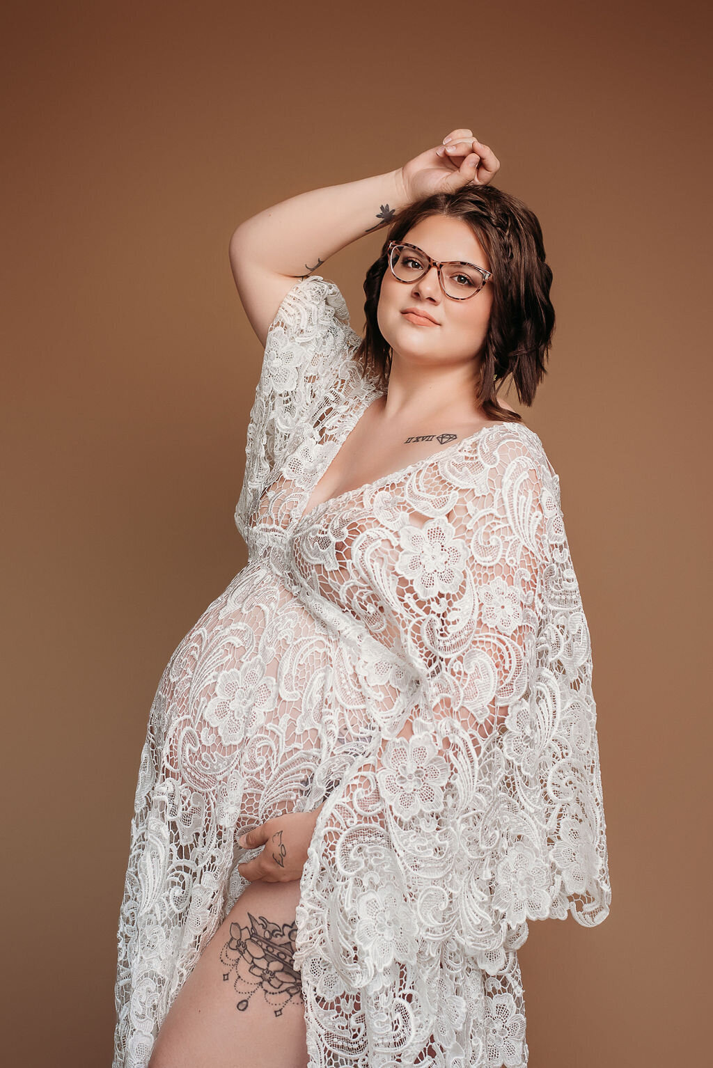 A pregnant woman in a white lace robe poses in front of a neutral-toned tan backdrop for her boho glamour maternity photoshoot, captured by a photographer in Bemidji, Minnesota