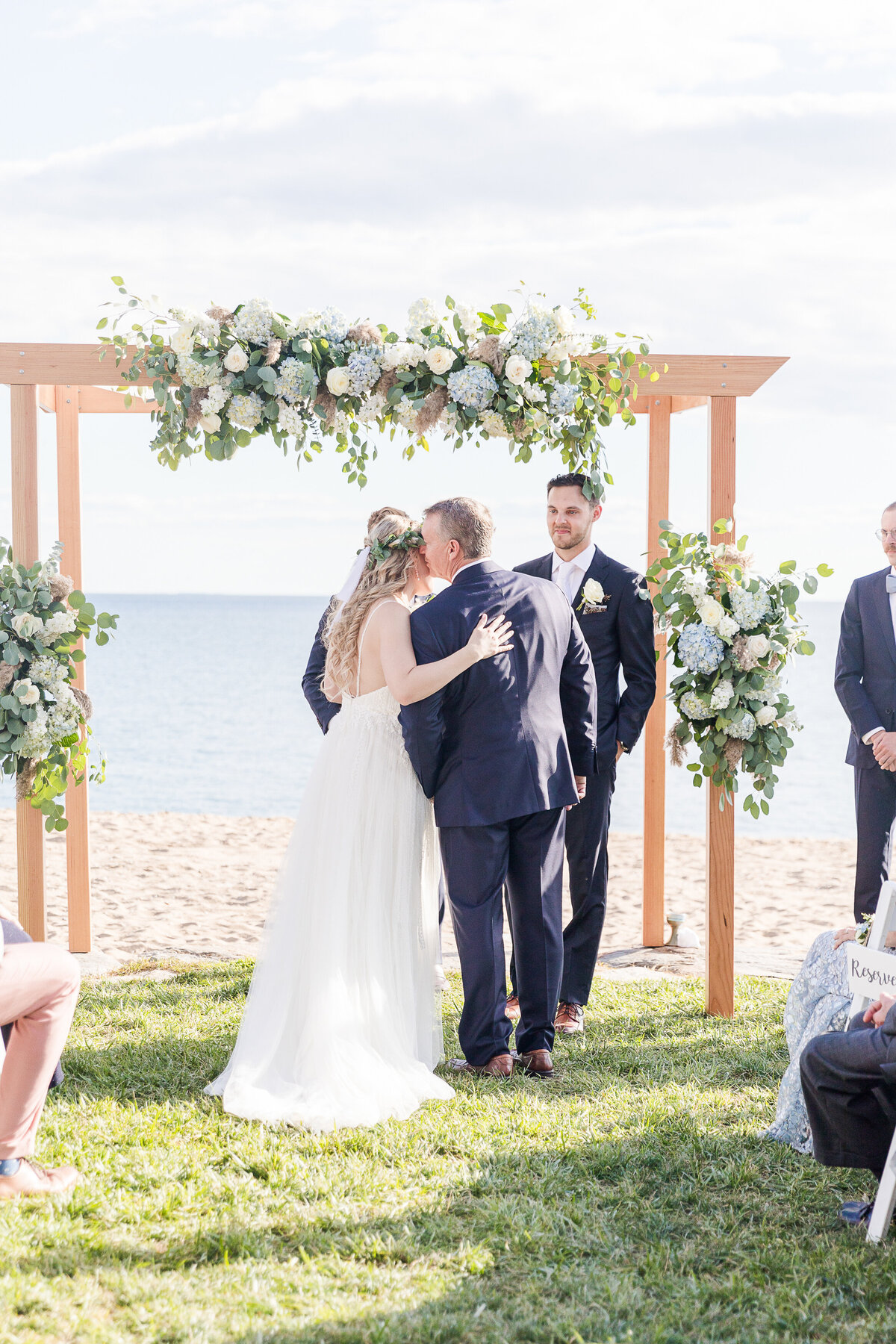 Father of the bride is seen kissing his daughter on the forehead as he gives her away at her wedding ceremony. Behind them is the ocean. Captured by best New England wedding photographer Lia Rose Weddings.