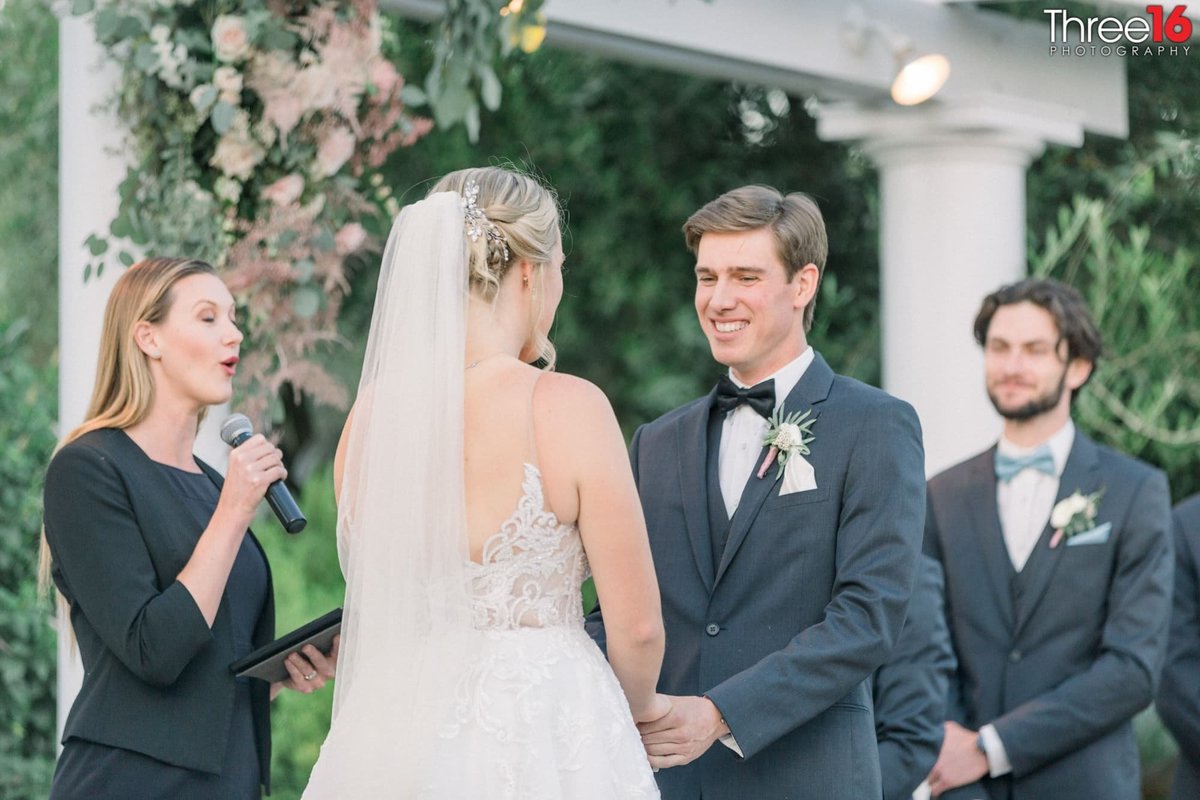 Groom smiles while holding his Bride's hands at the altar as the officiant is speaking