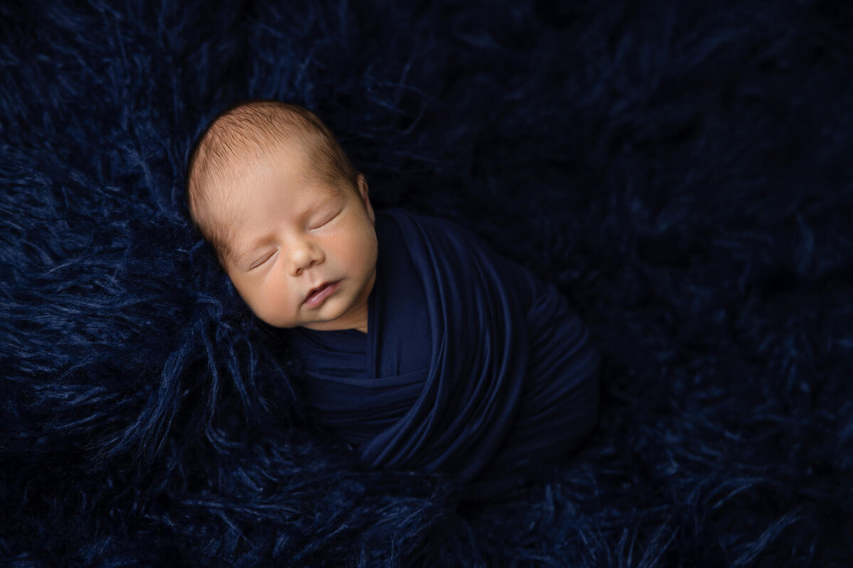 newborn baby asleep on a blue shaggy rug wrapped in blue fabric