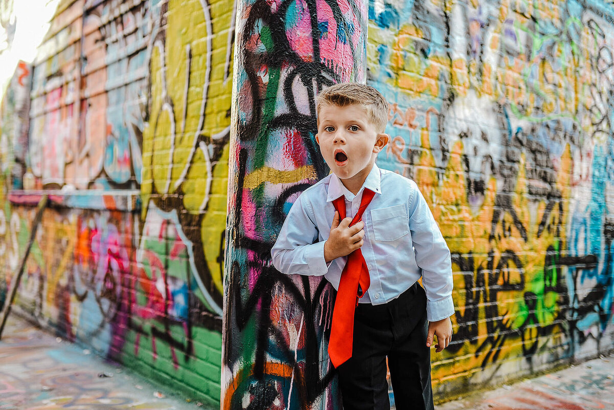 Little boy in a white shirt and black pants pulling off a red tie in graffiti alley near MICA in Baltimore Maryland