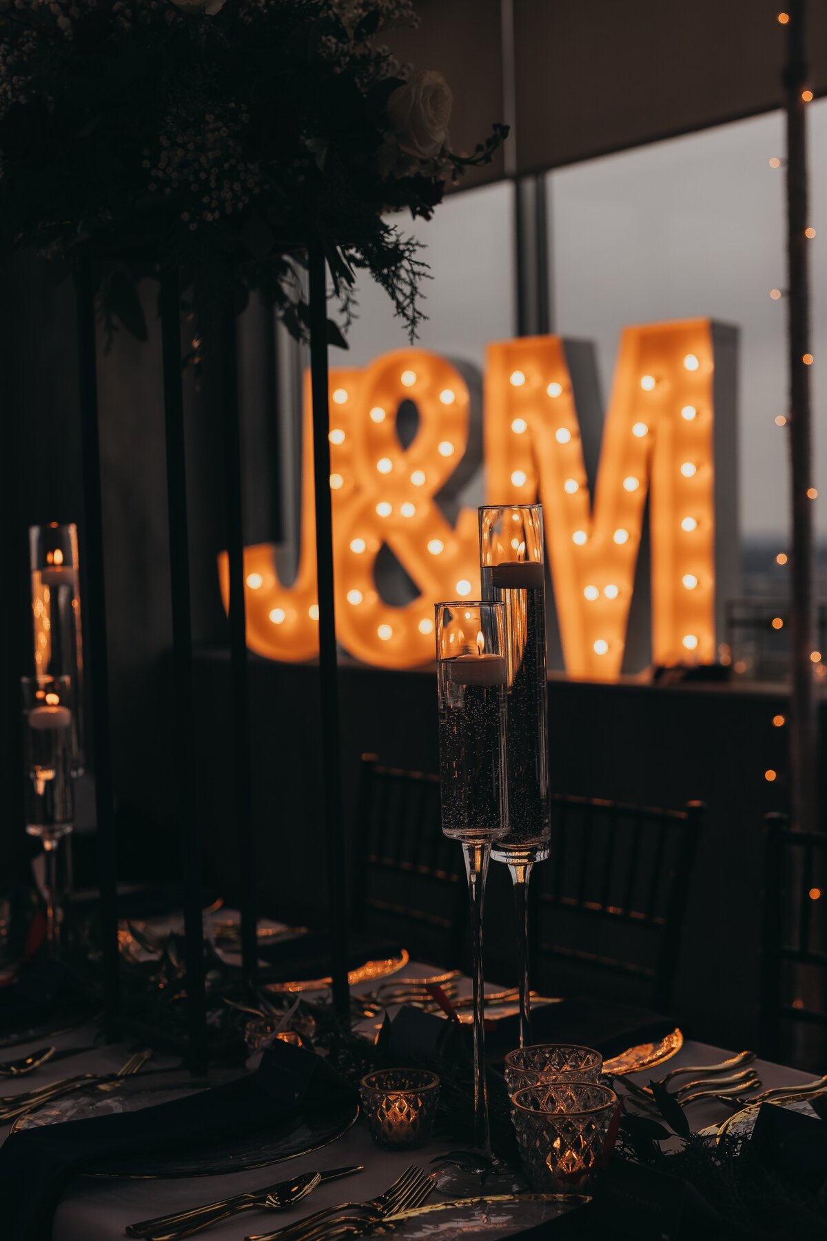 Elegant dinner table setting with champagne flutes, lit candles, and large illuminated letters "J&amp;M" in the background at dusk, expertly organized by a top wedding coordinator from Des Moines.