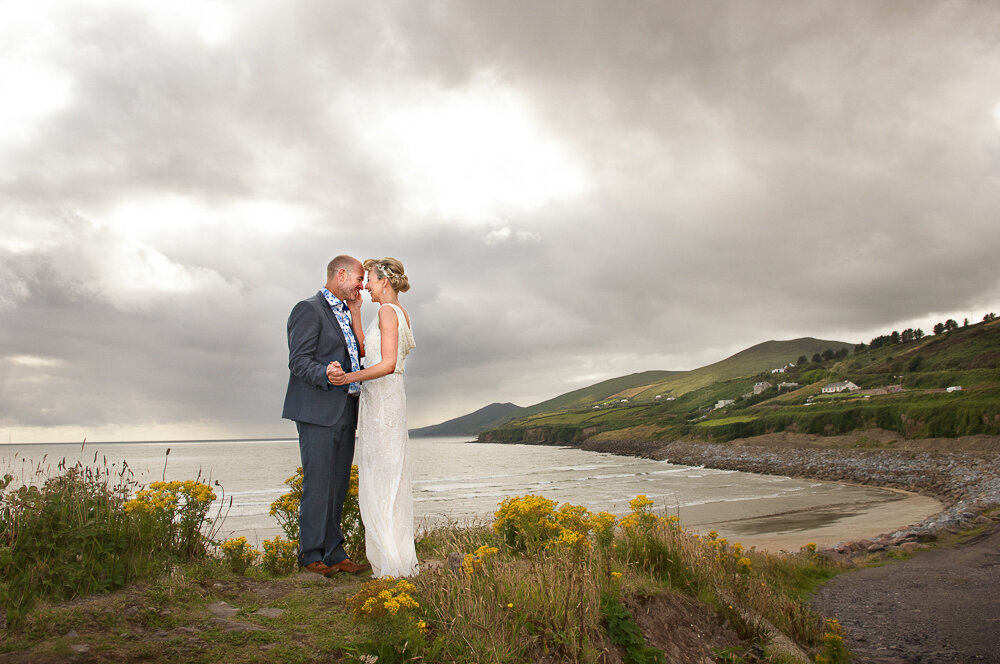blonde bride wearing a scoop -back, column style dress embracing her groom, wearing a blue suit and floral shirt standing on the edge of Inch beach, Kerry