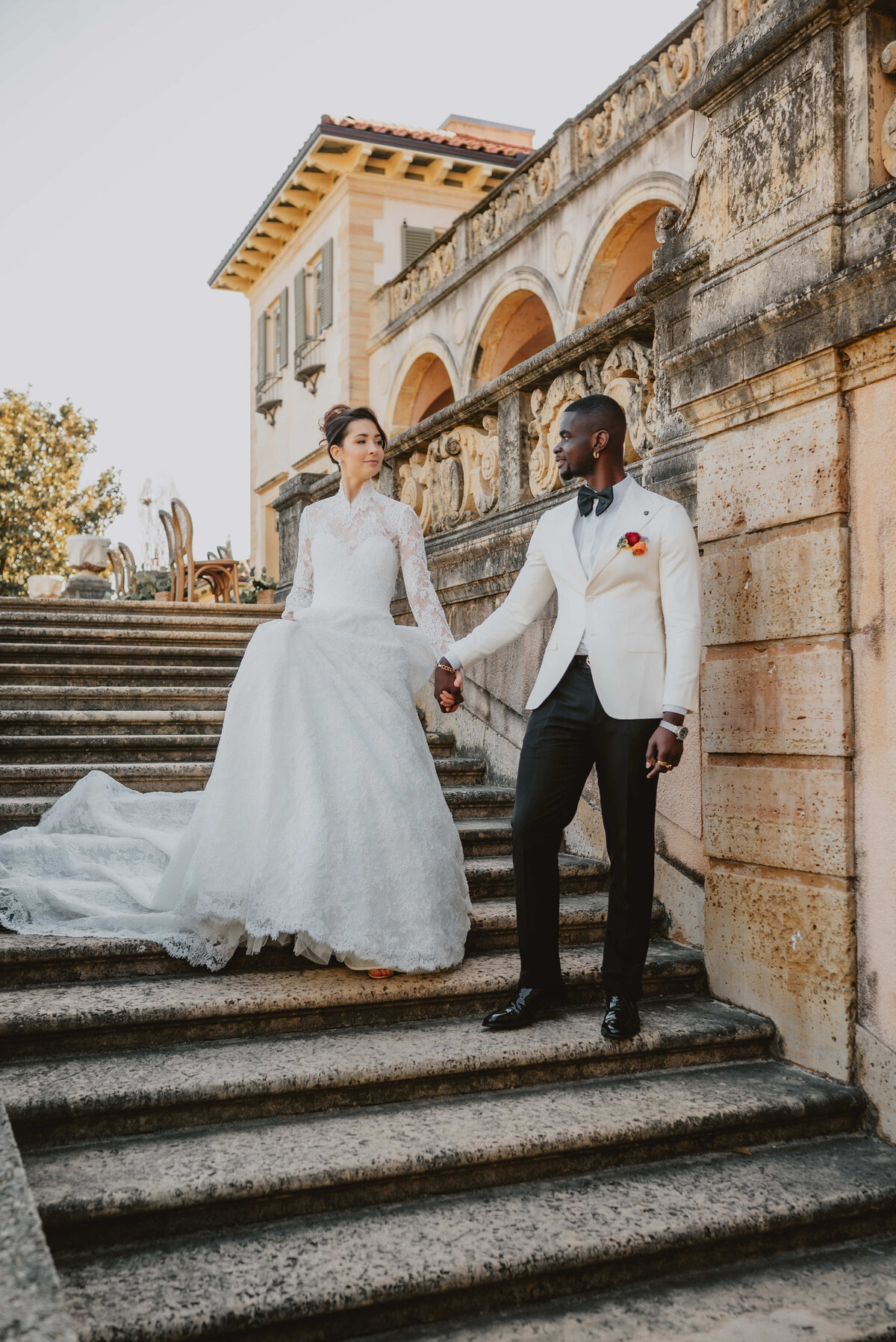 A bride in a white gown and a groom in a white suit with a red boutonniere hold hands, walking down ornate stone steps, exuding elegance and joy.
