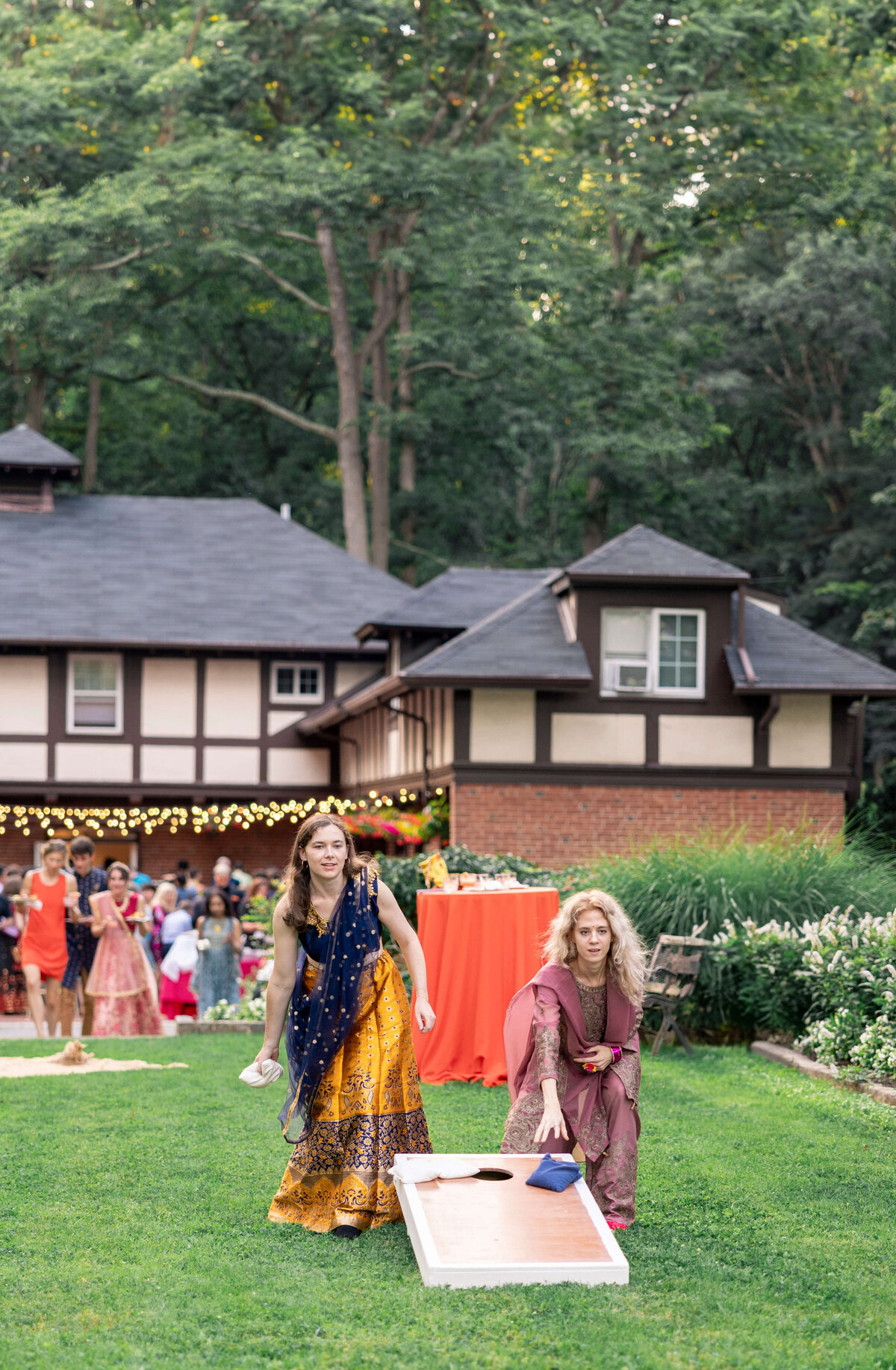 Two women in colorful traditional attire play a game of cornhole on a grassy lawn. A group of people and a Tudor-style house are in the background, with lights strung across the scene, suggesting a festive outdoor gathering.