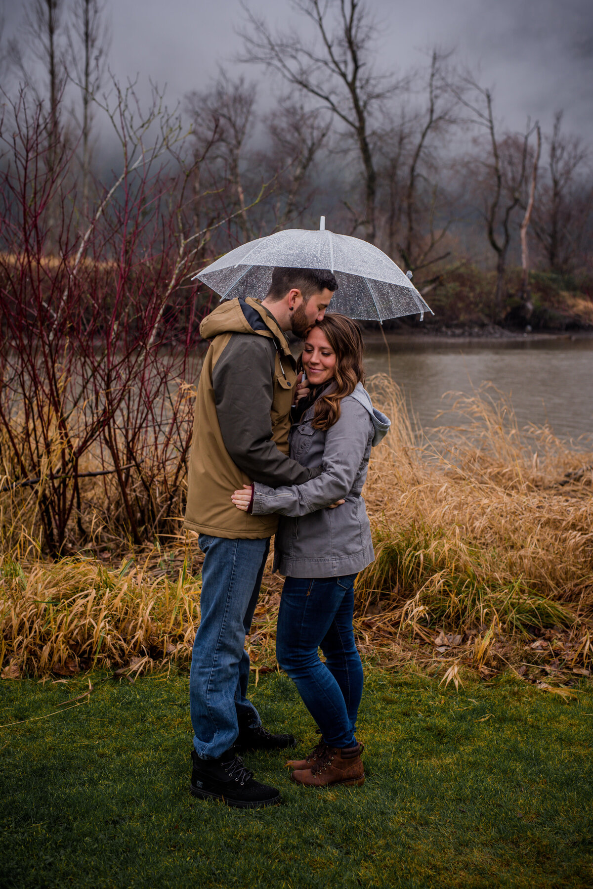 Rainy Abbotsford BC engagement. Couple stands under an umbrella, guy kisses girl forehead and girl eyes are closed. In the back round has a lake and trees with no leaves. 