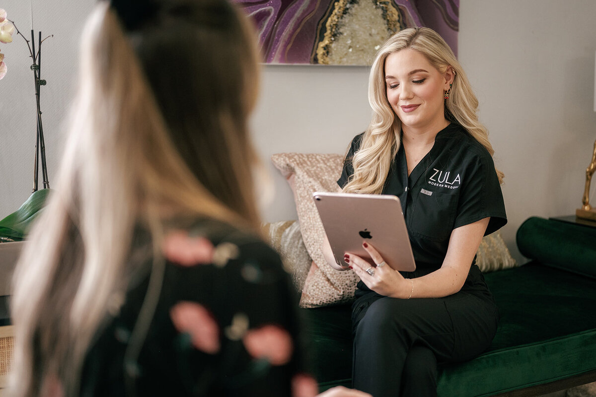 Medspa practitioner sitting on a green couch working on an ipad talking to a patient