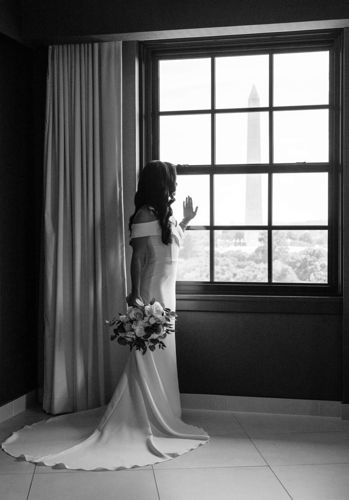 A bride in an off-the-shoulder gown stands by a window, holding a bouquet and gazing out at the Washington Monument. The room is elegantly lit, and the scene captures a moment of reflection.