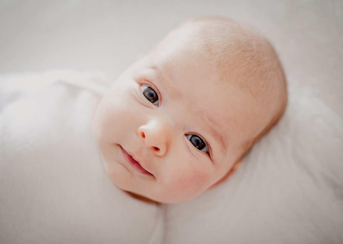 Baby looks straight to camera in a mesmerizing portrait. She is wearing a plain white onesie and laying on a white bedspread.