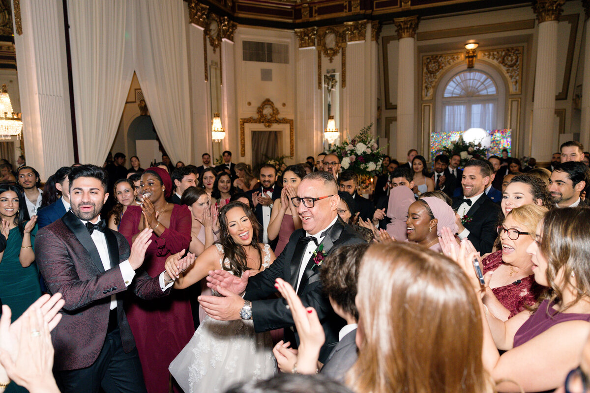 A joyful wedding celebration with a bride and groom dancing surrounded by guests in elegant attire. A beautifully decorated hall serves as the backdrop, with ornate details and a chandelier visible.