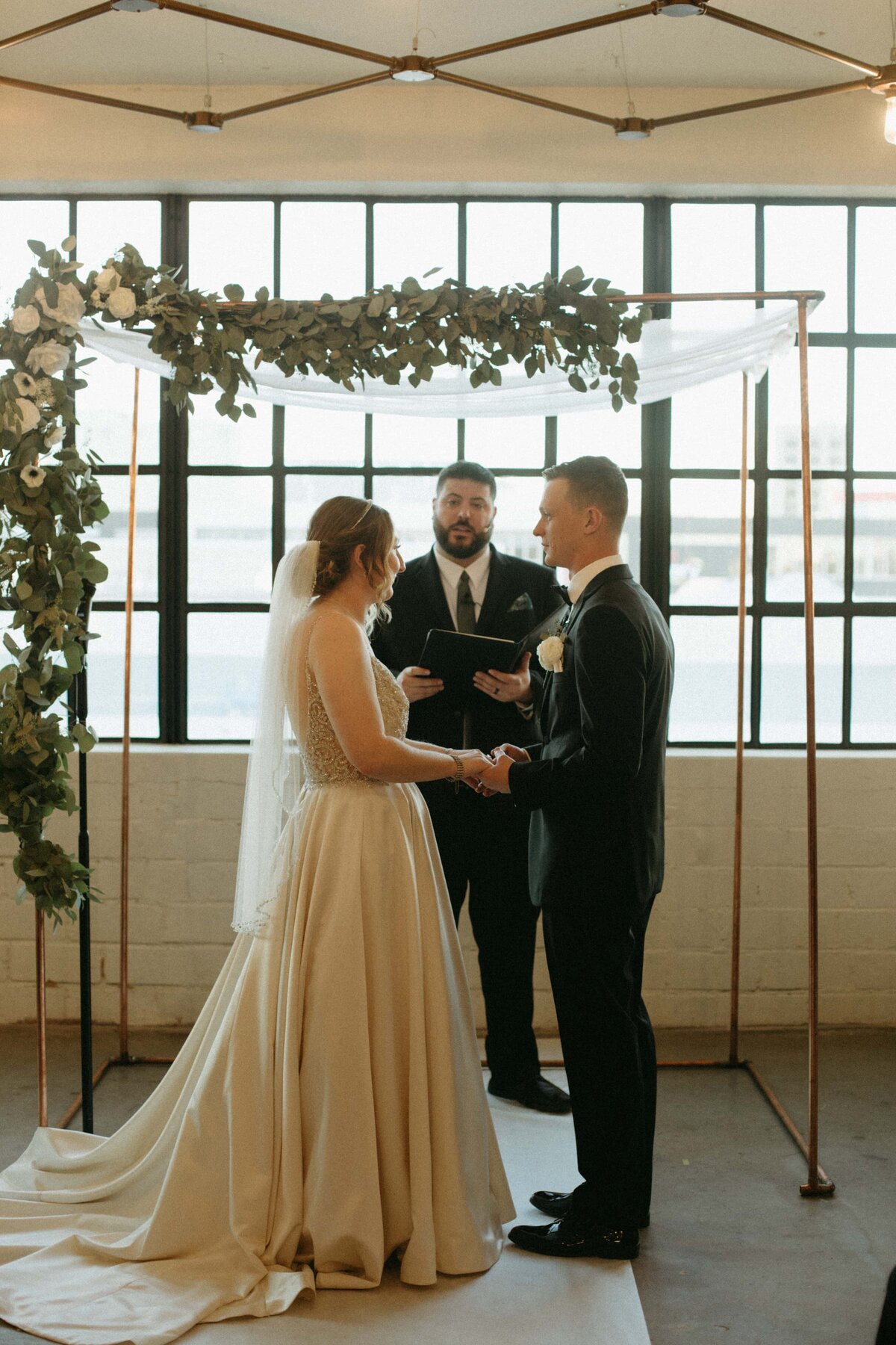 Bride and groom exchanging vows in front of an officiant under a floral arch inside a bright room with industrial windows at an Iowa wedding.