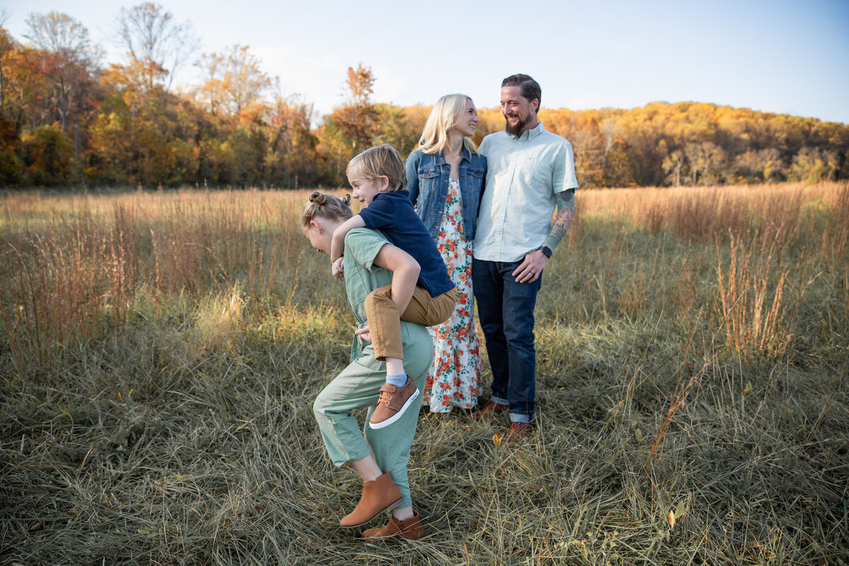 A brother rides on his sister's back while the baltimore county parents stand in the distance smiling at eachother during their lifestyle family session