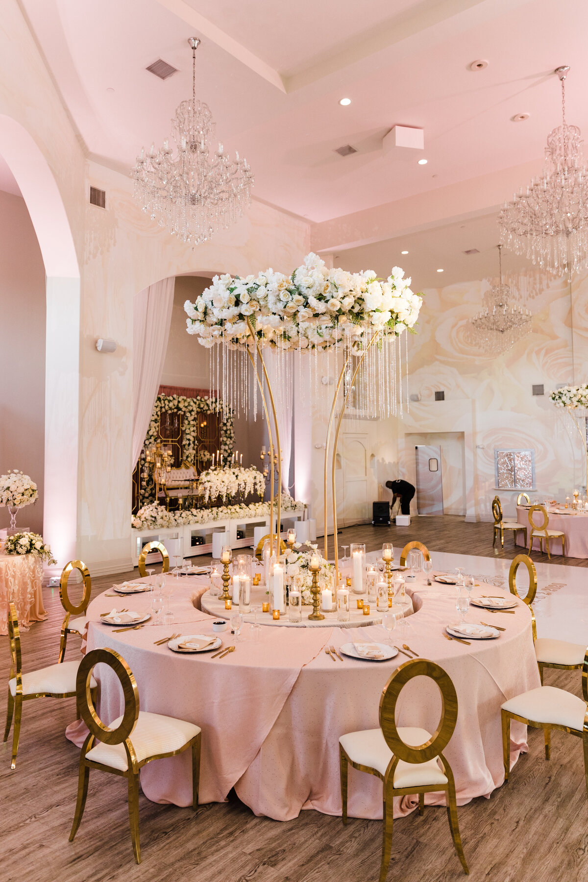 A detail shot of a table and room for a wedding reception in DFW, Texas.  The large, round table is covered in a light pink tablecloth, place settings, many candles of different shapes and sizes, and a large, tall, intricate floral centerpiece that towers above the table itself. The room is white and light pink with more reception tables, multiple chandeliers, a dance floor, and a lavish head table adorned with lots of floral arrangements.