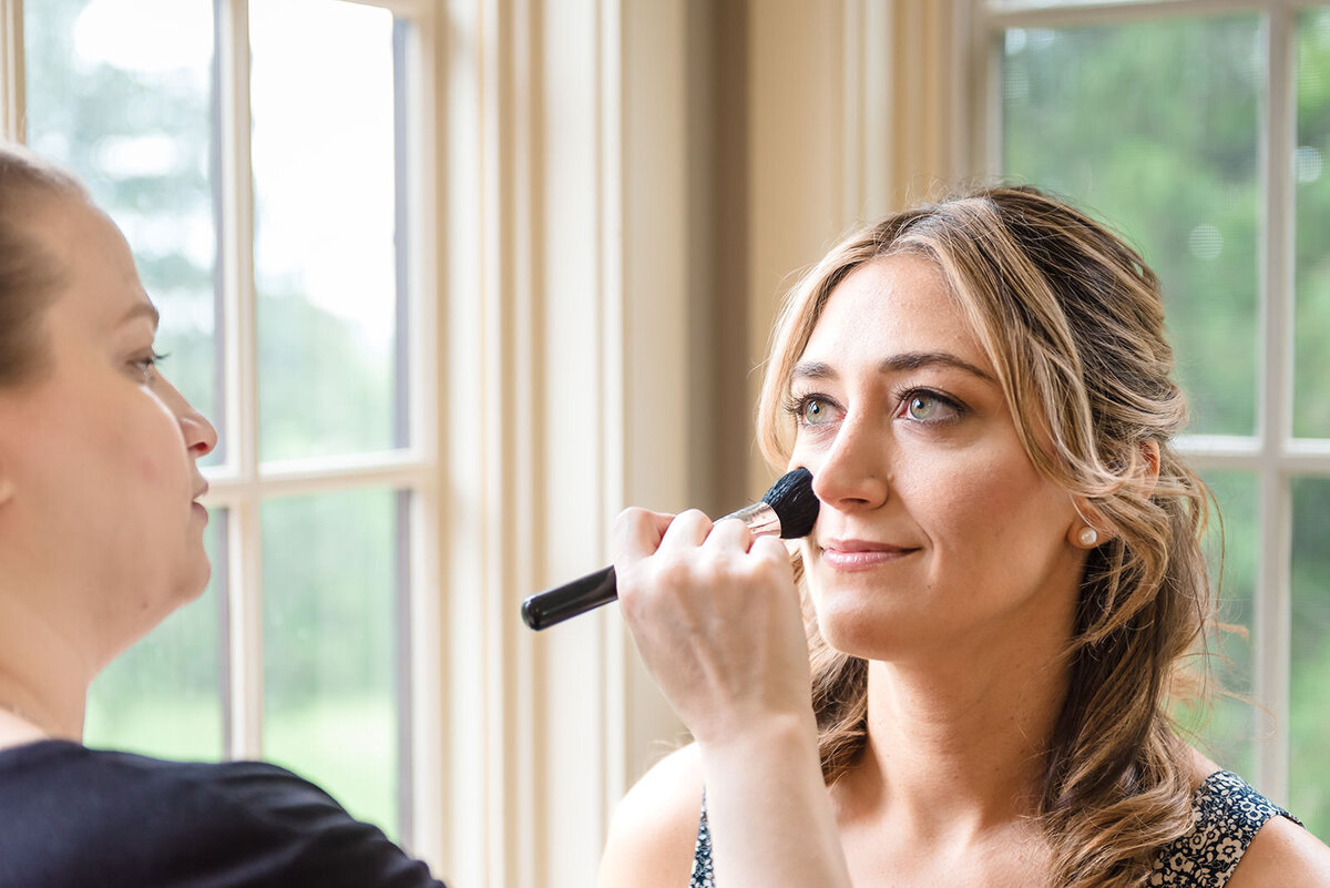 A woman having makeup applied with a brush by another person in a room with a window view.