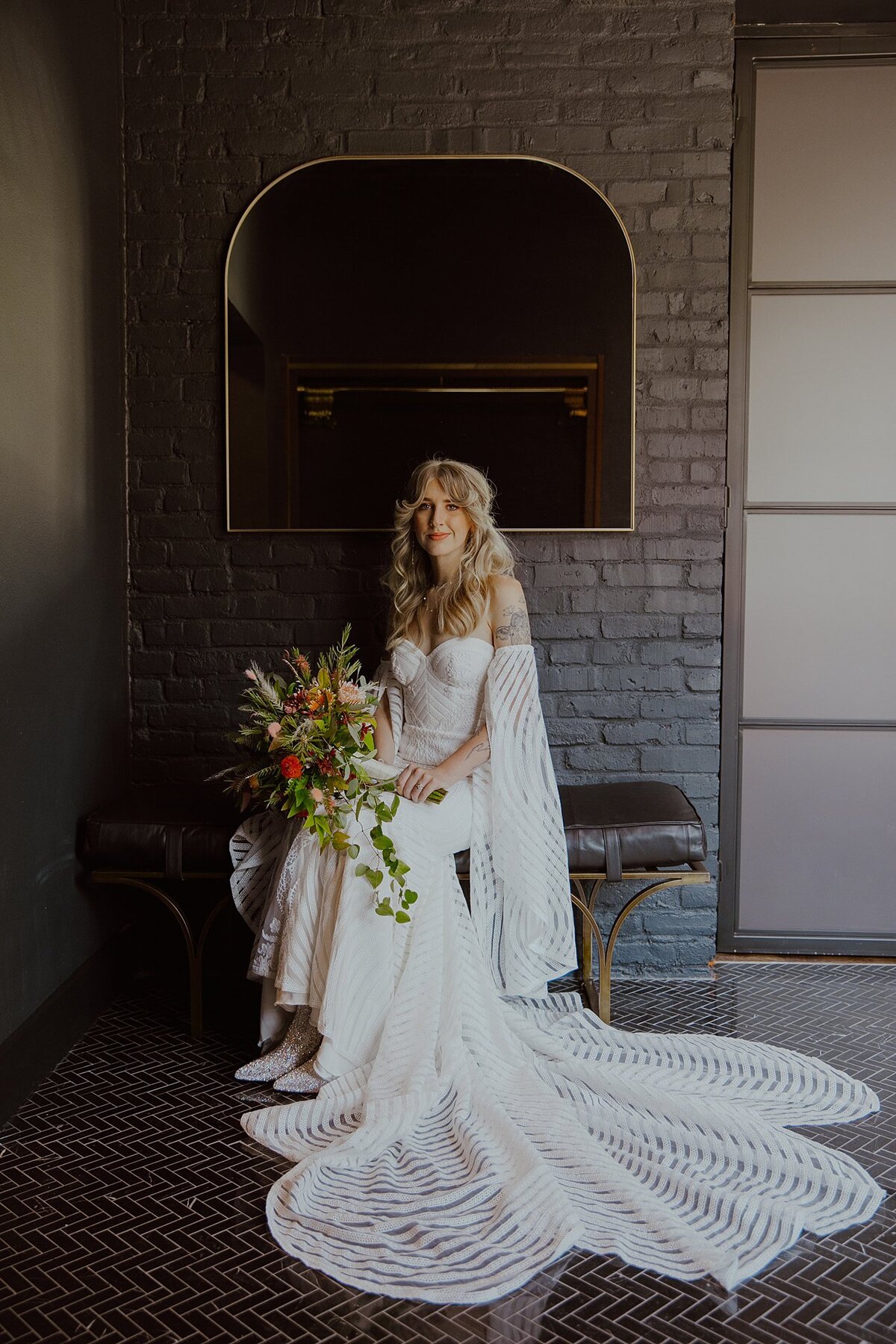 Boho bride in a strapless striped wedding dress with detached bell sleeves wearing white cowboy boots sits on a brass bench with a brown leather cushion holding a large cascade bouquet of orange, red and yellow flowers with a black herringbone tile floor in the foreground and exposed brown brick behind her at Clementine Nashville.
