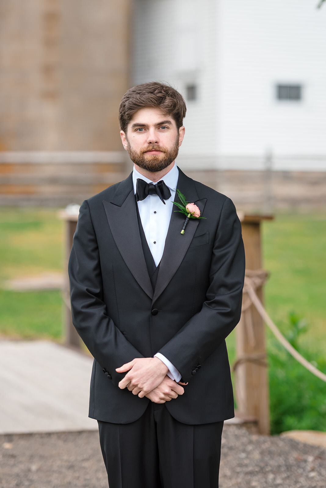 A groom in a black tuxedo standing outdoors with his hands crossed in front of him.