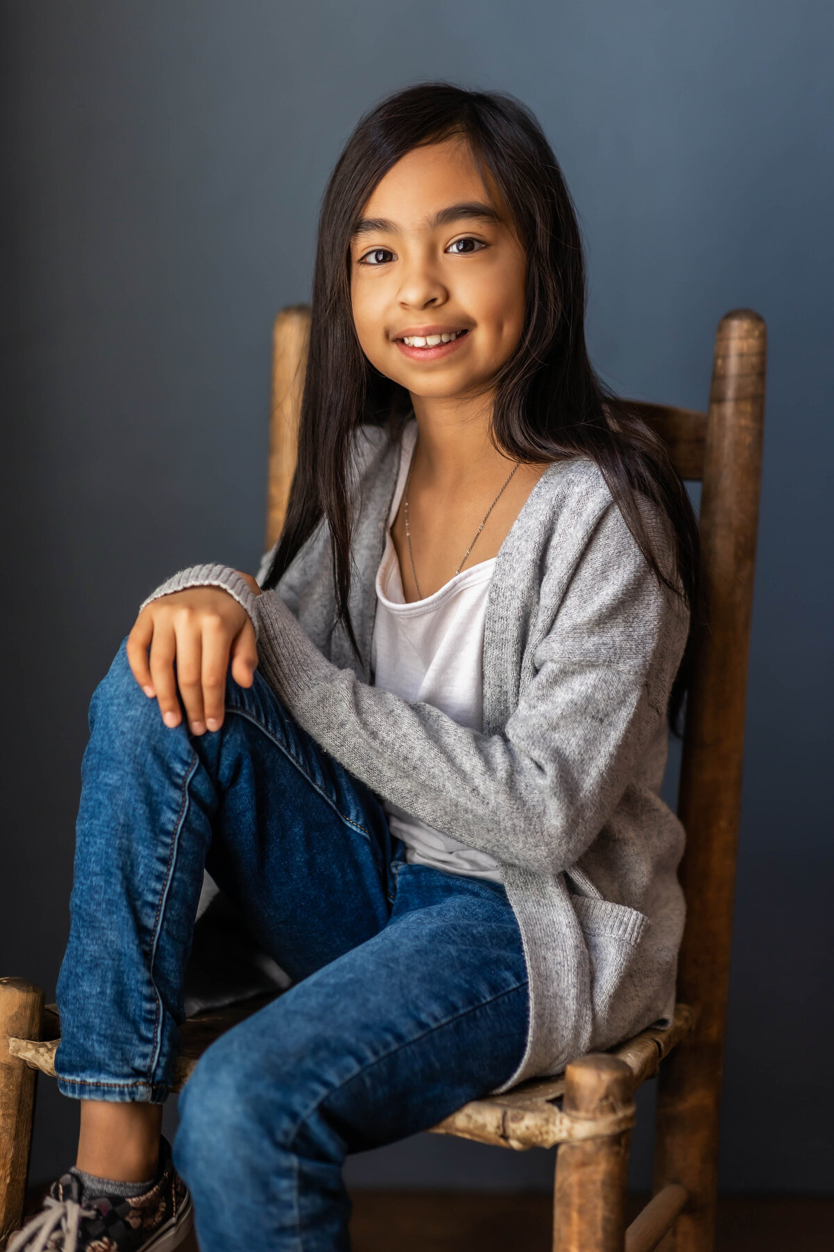 Young girl is wearing a gray sweater with white tshirt and jeans and is seated and smiling at the camera at an indoor session