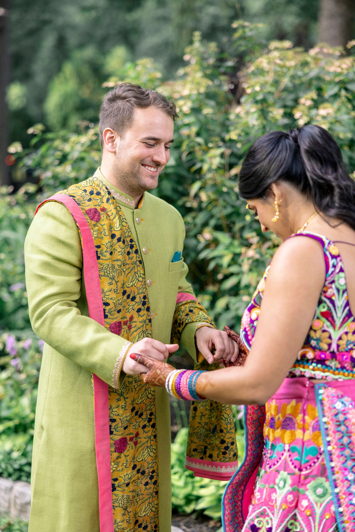 A person in a green and gold embroidered sherwani is smiling and holding hands with another person in colorful traditional attire, adorned with bangles. They stand outdoors surrounded by greenery.
