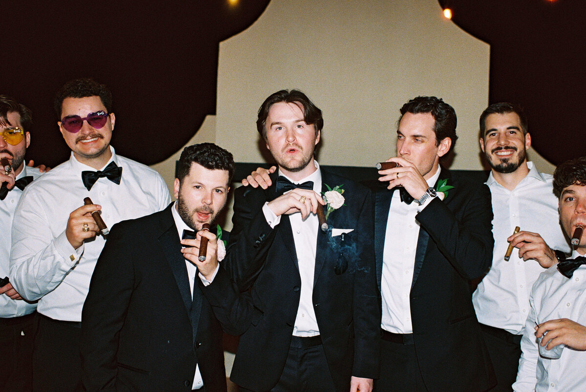 A group of seven men in tuxedos smile and enjoy cigars at a celebratory event. Festive atmosphere with dim lighting, exuding camaraderie and joy.