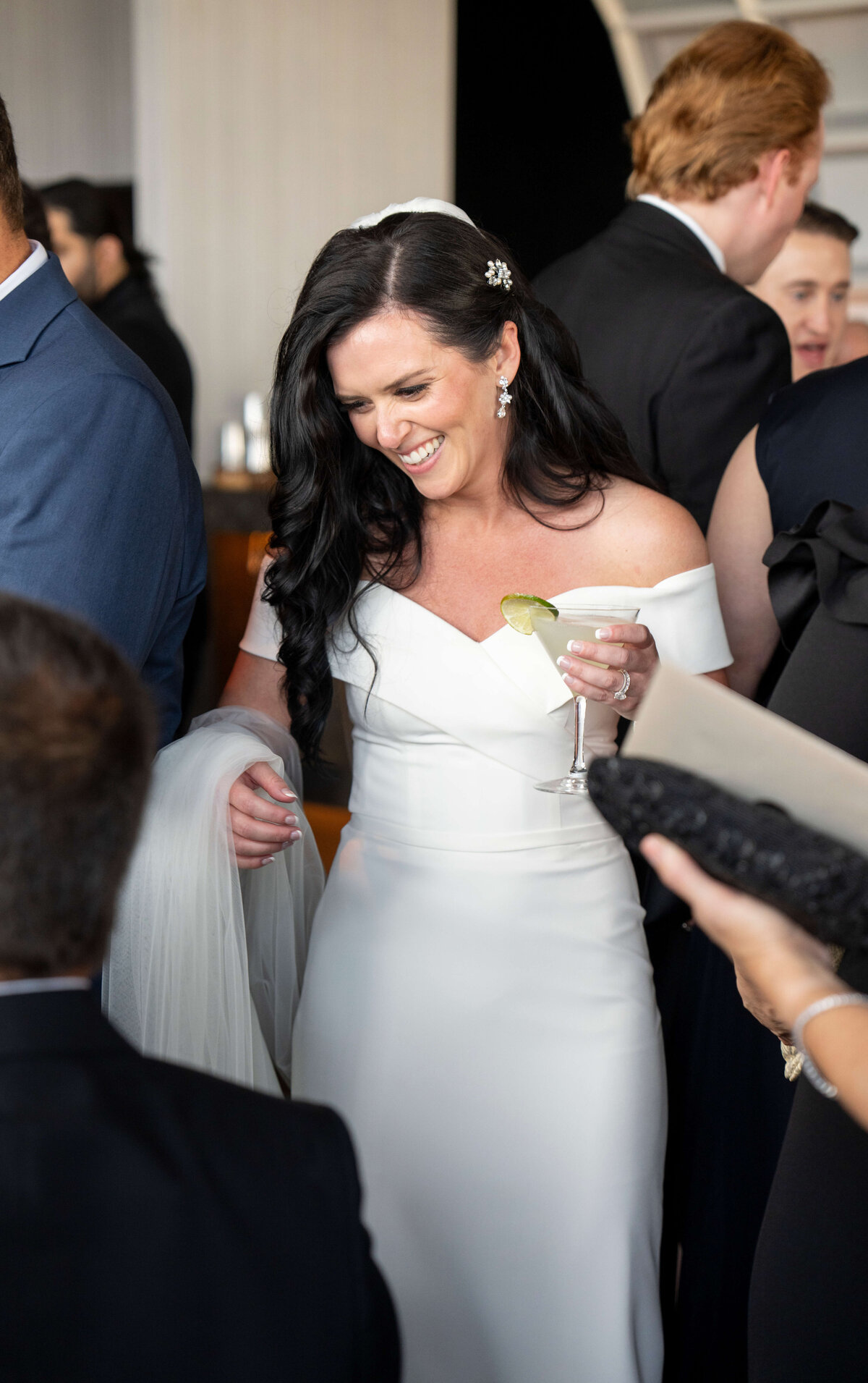 A smiling woman in an off-the-shoulder white dress holds a cocktail with a lime garnish. She stands among people at an event, draped in a white shawl. Her dark hair features jeweled hairpins, and she wears earrings.