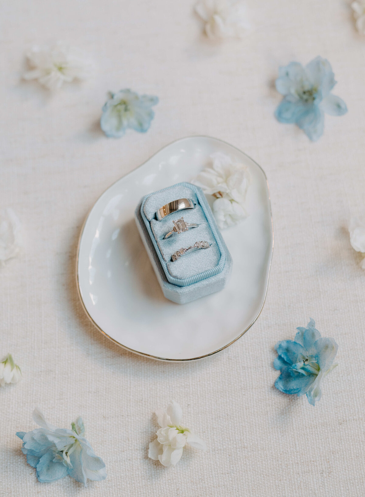 A pale blue ring box with three rings, placed on a white ceramic dish, surrounded by delicate white and blue flowers on a soft beige background.