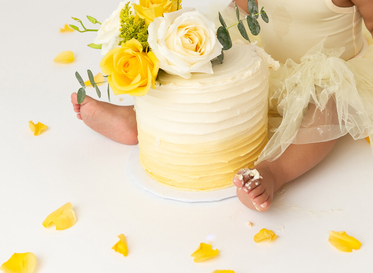 Baby girl in yellow tutu sits with yellow ombre cake topped with yellow roses between her legs. Close up image of cake and icing on her toes.