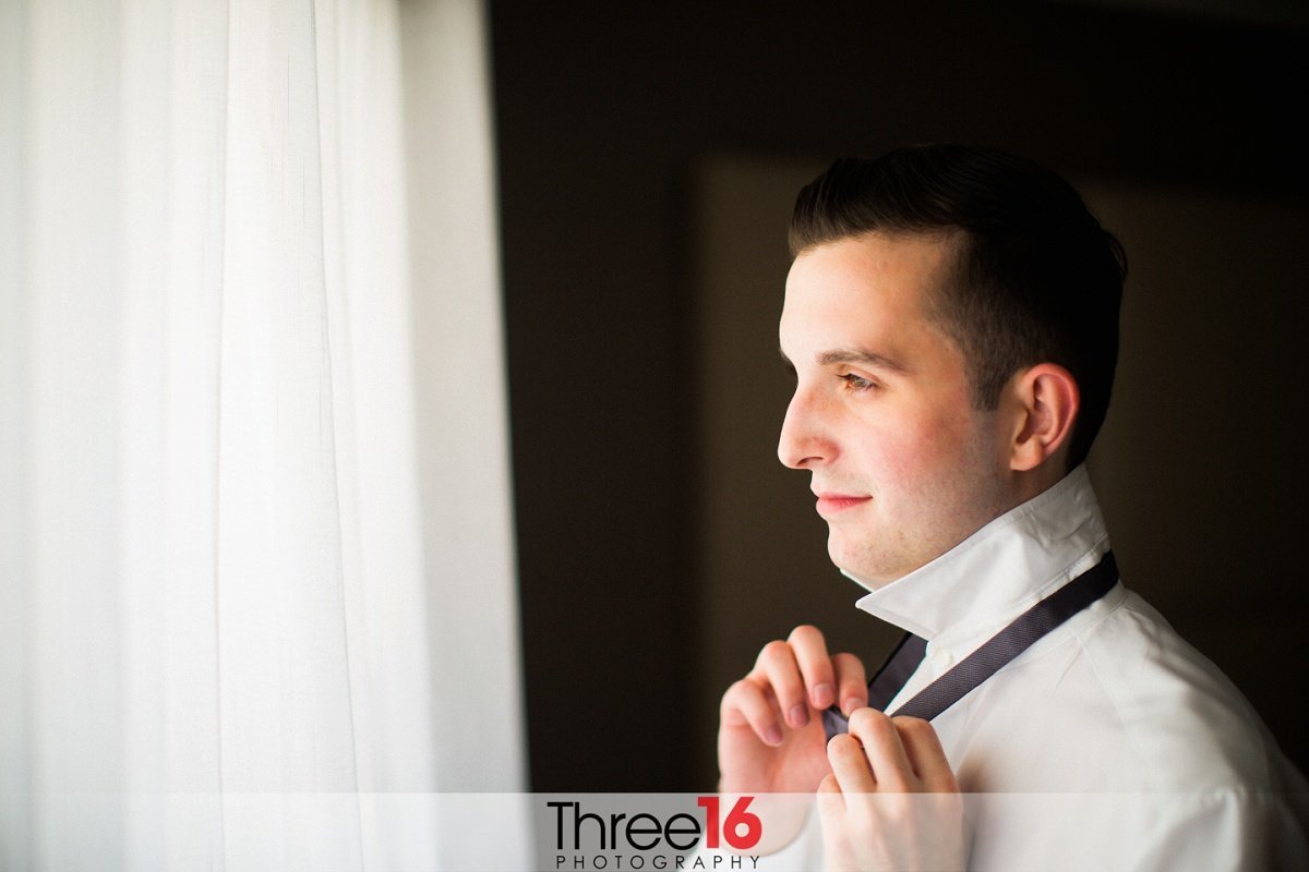 Groom puts on his bowtie as he looks out the window