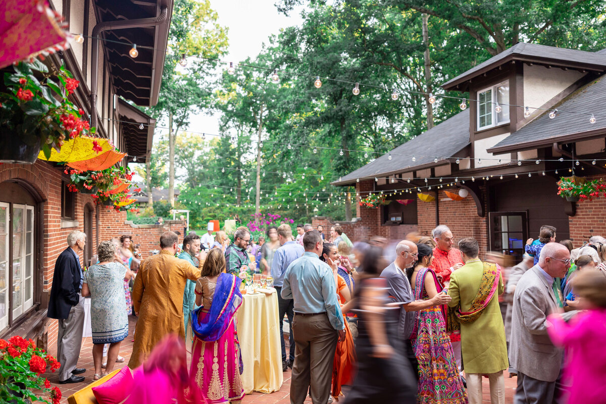A vibrant outdoor gathering with people in colorful traditional attire. Guests are mingling in a courtyard adorned with flowers and string lights. The setting is a charming brick building surrounded by greenery.