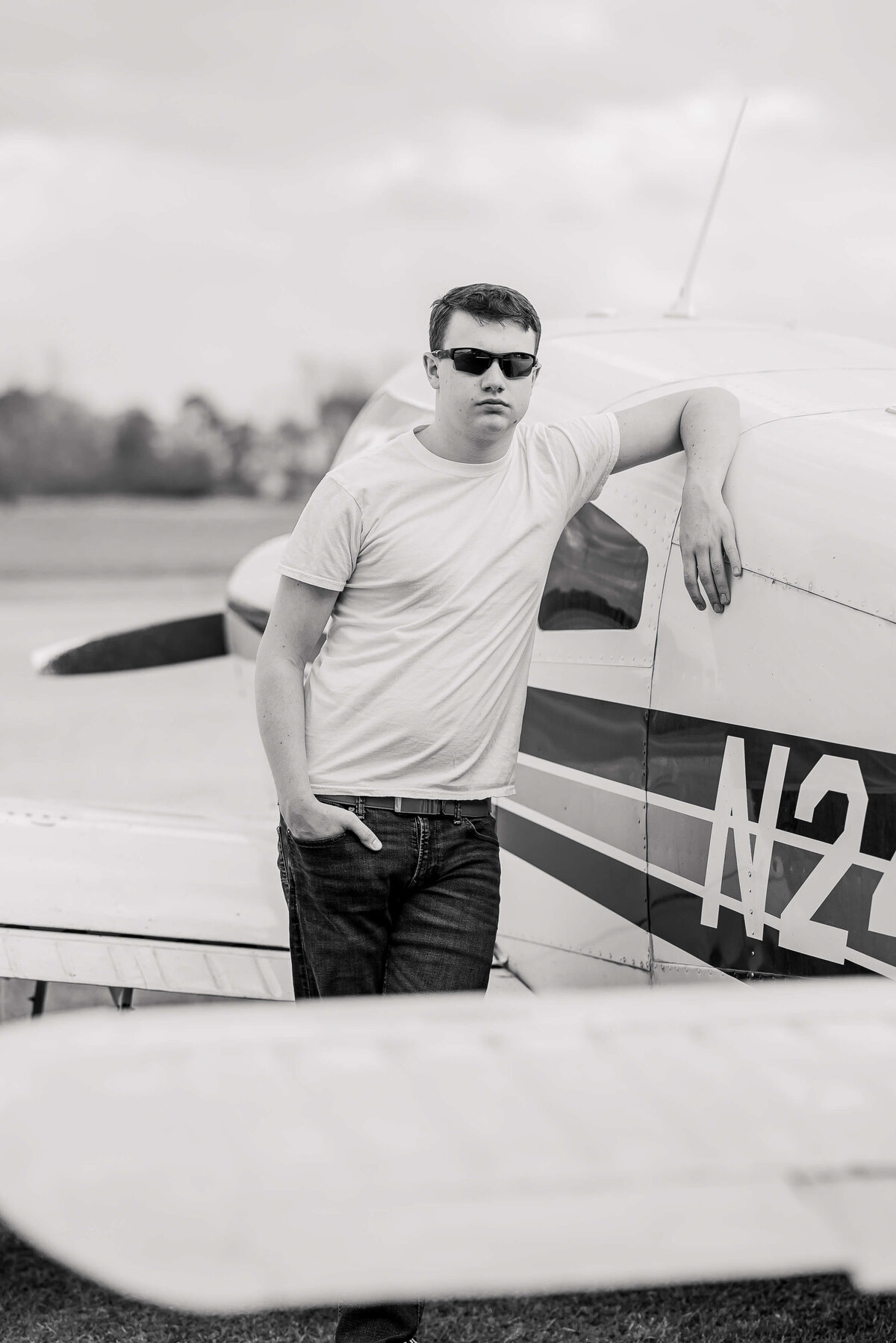 A man wearing sunglasses and a white t-shirt is leaning casually against a small aircraft. The plane, featuring part of a visible registration number, is parked outdoors on an airfield. The image is in black and white.