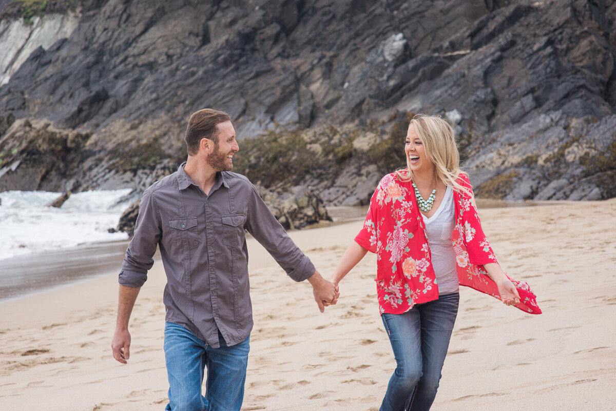 Young couple holding hands and wearing denim, laughing while running on the beach in Kerry