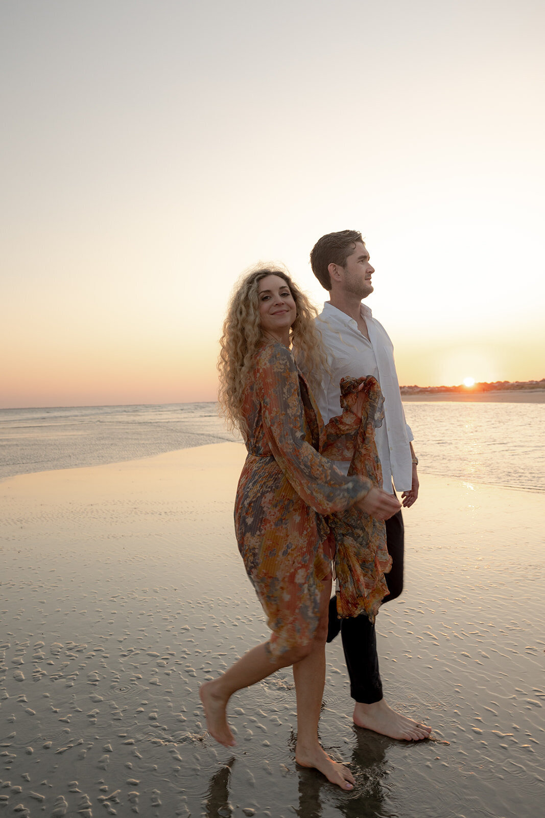 Couple walking side-by-side at the bech. Woman in orange dress with flower pattern is walking in foreground and holding on to man's arm. Sun is setting in the background and reflecting romatically on water.