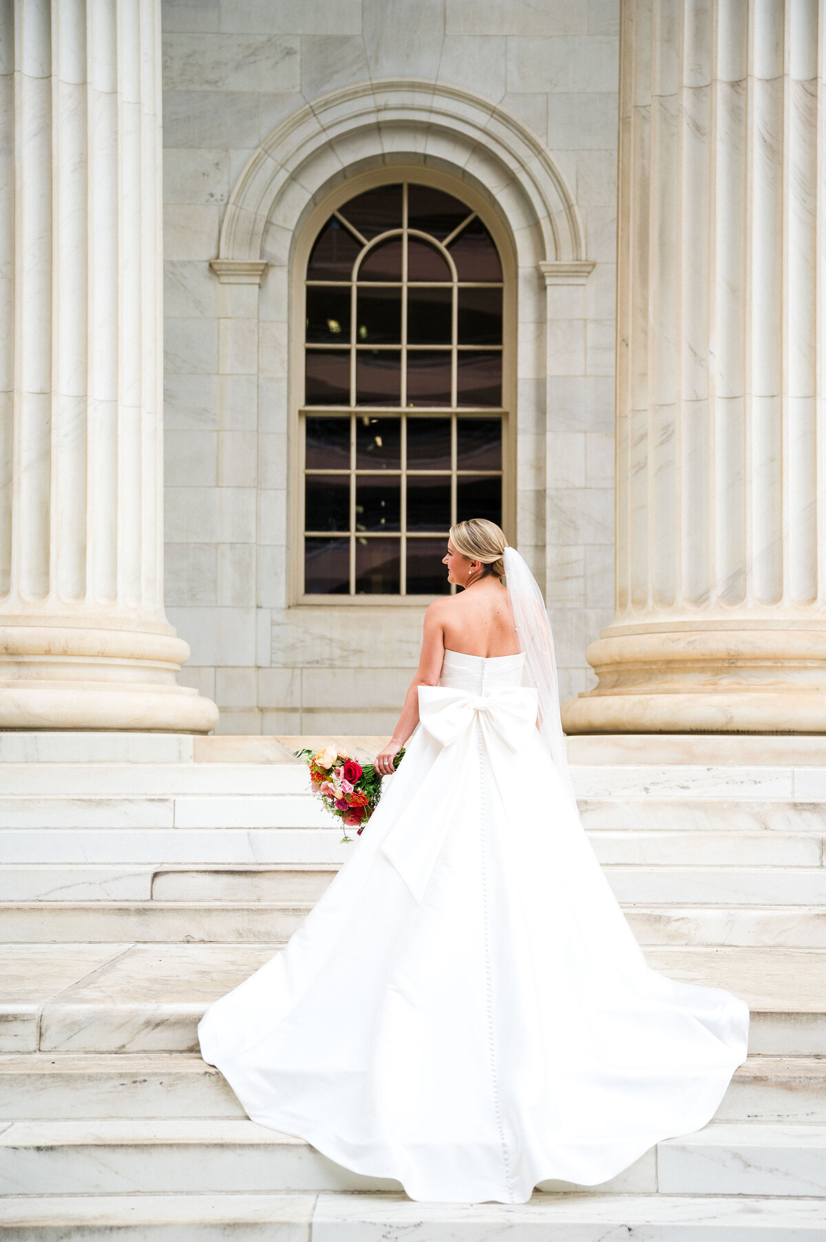 A bride faces away from the camera and the train of her wedding gown is draped over the stairs of a downtown Denver building.