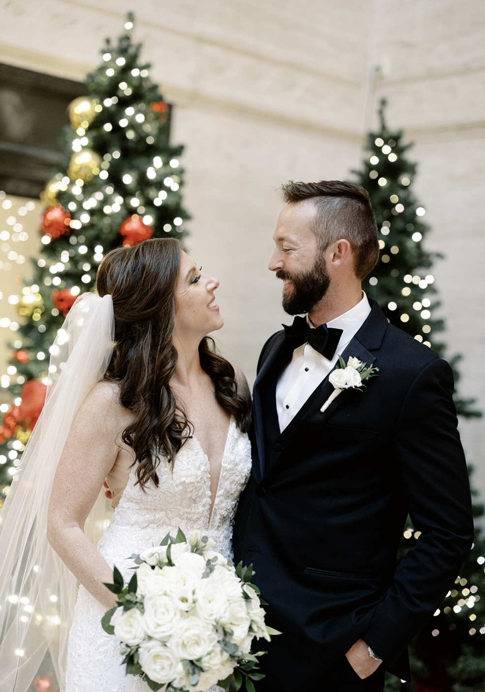 Bride with brown curly hair holds bouquet of white roses and smiles at husband wearing a black tuxedo in front of Chirstmas trees at Harold Washington Library wedding reception.