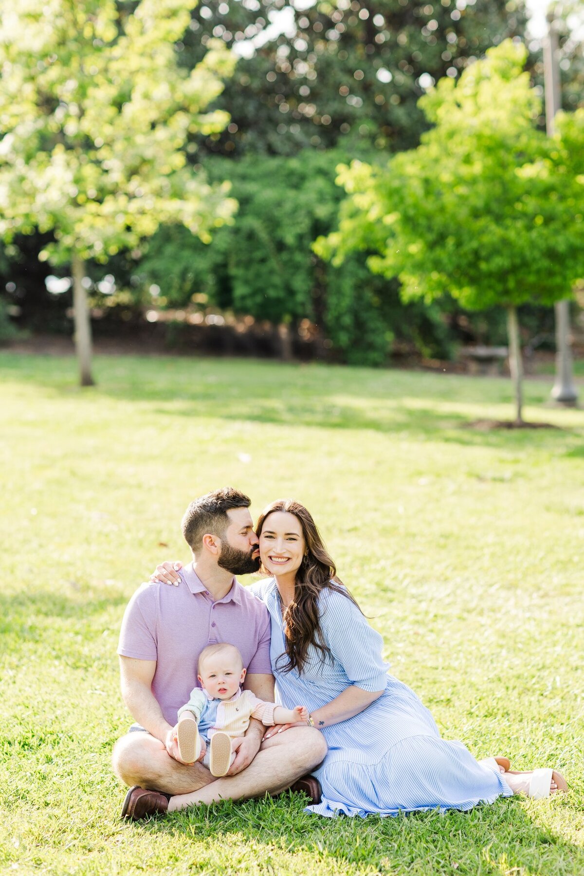 Husband kissing wife on the cheek while baby boy looks at the camera and smiles in a green field during spring
