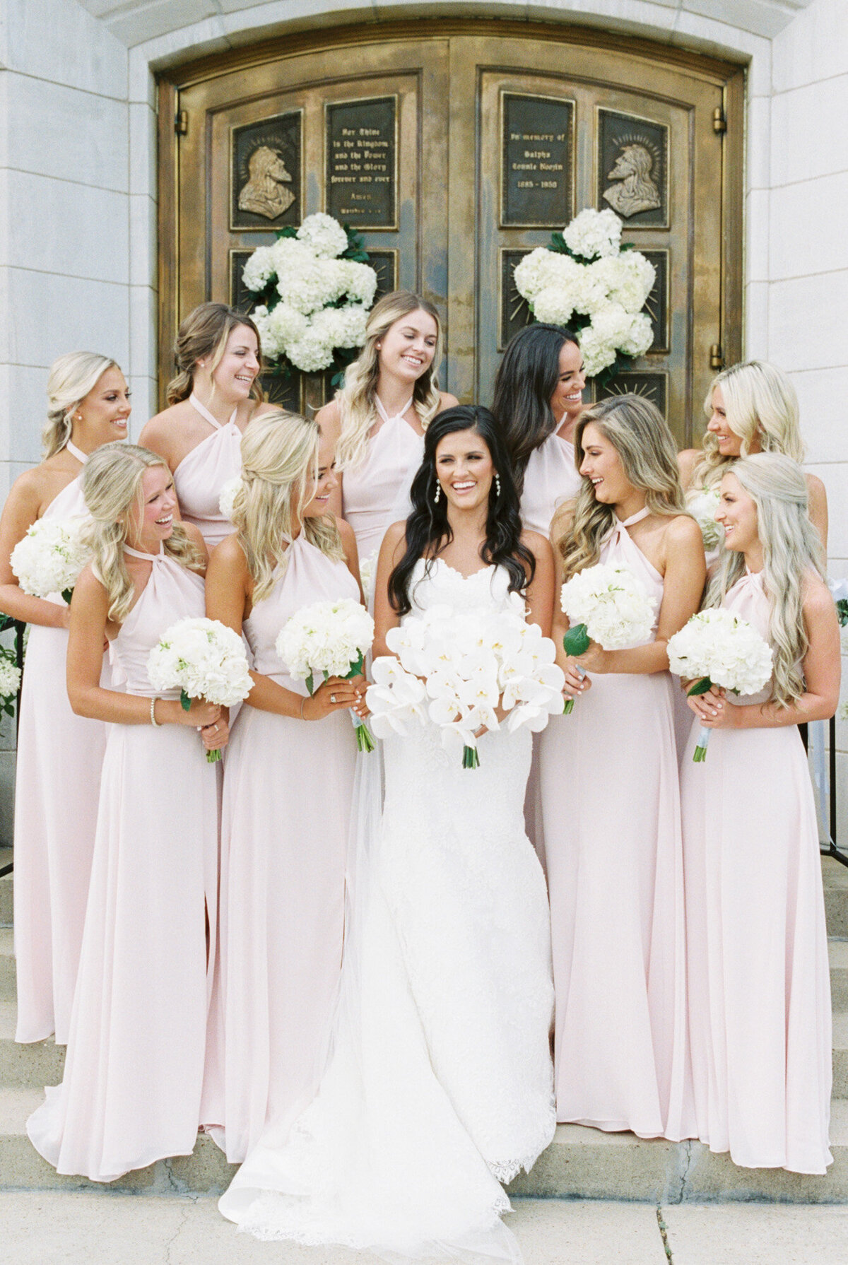A bride and her bridesmaids stand in front of the church door as they laugh by Birmingham wedding photographer, Kelsey Dawn Photography