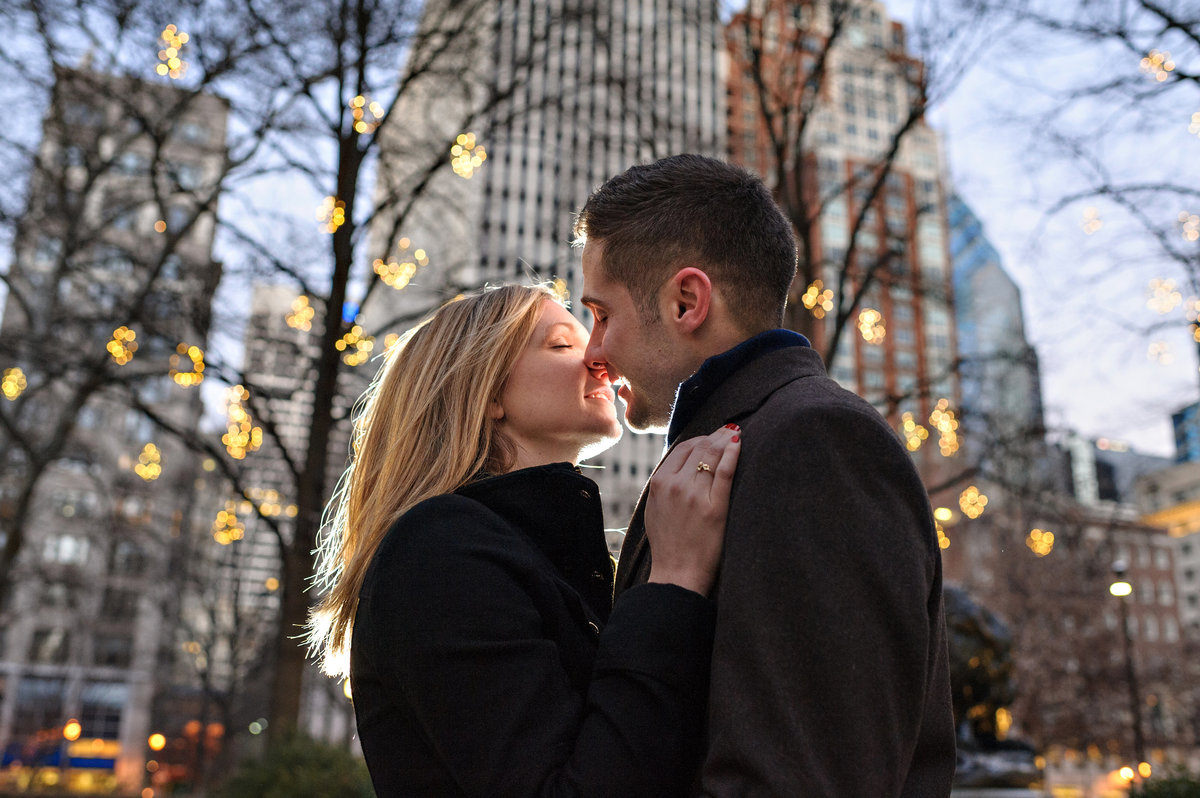 Surrounded by holiday lights in rittenhouse square, an engaged couple share a kiss.