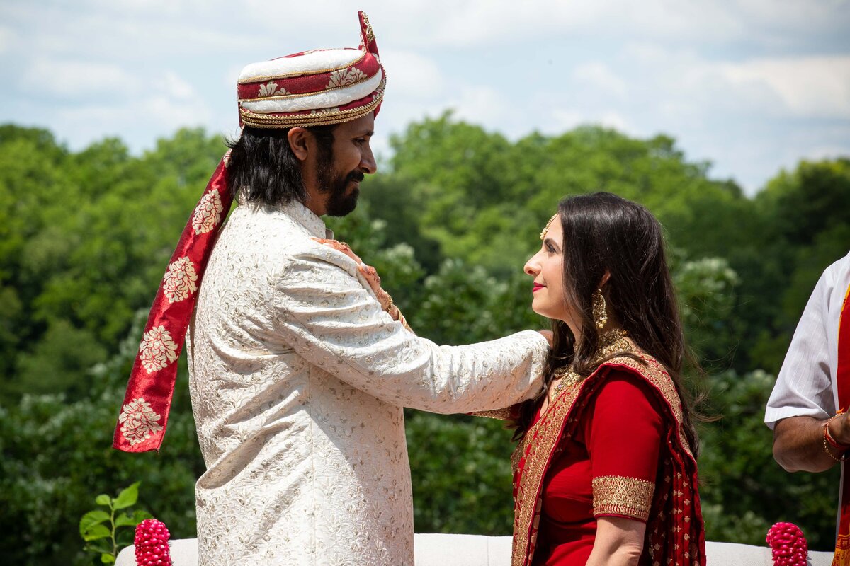 A bride in a red saree and a groom in a white sherwani with a red turban share a tender moment outdoors at an event in Davenport.