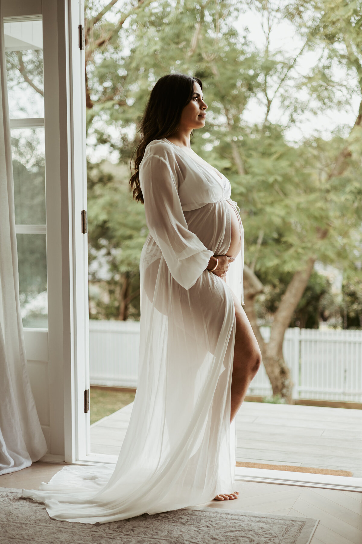 Pregnant woman in white see-thorough robe posing in doorway of French doors