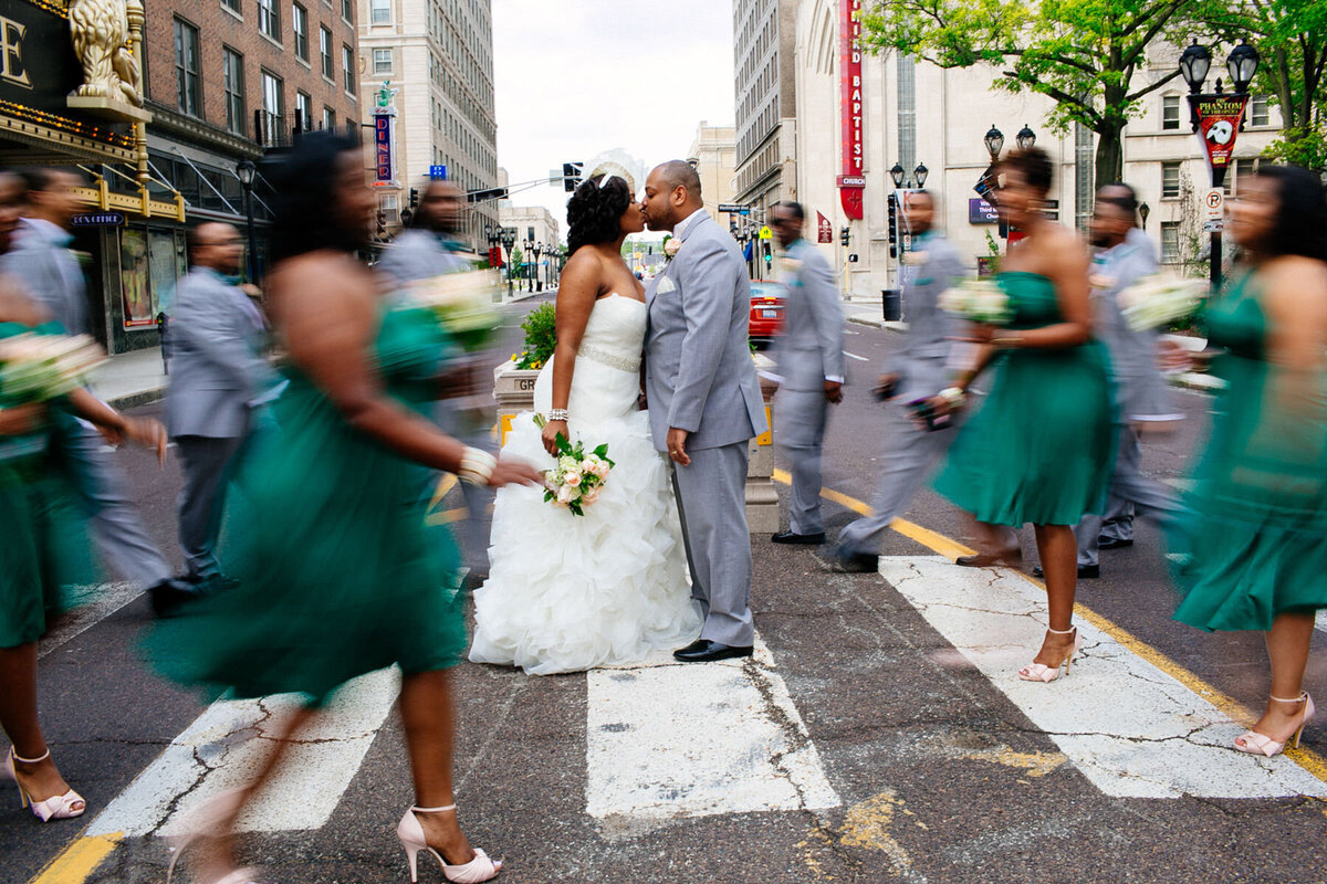 Bridal party members cross the street, blurred from movement, as the bride and groom hold still in downtown St Louis.