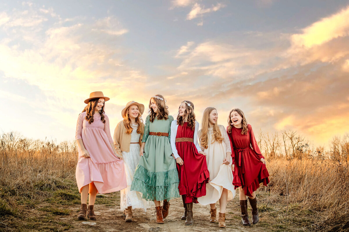 6 teenage girls in colorful dresses walking together side by side and laughing in a field at sunset