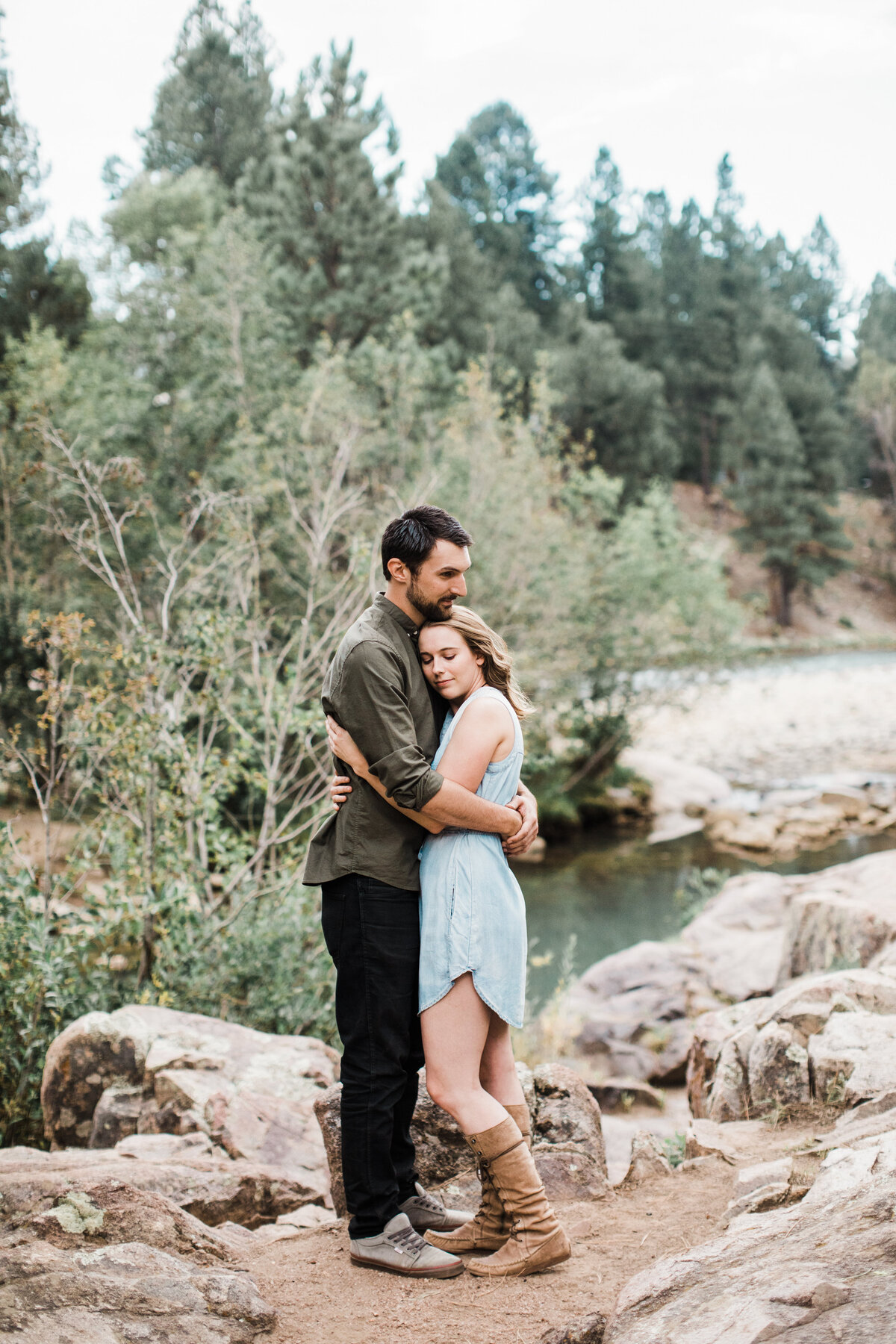 A couple holding each other close in front of the Animas River during their engagement session in Durango, Colorado. The woman on the right is wearing a short blue dress and boots while the man on the left is wearing a dark green dress shirt and dark pants. Behind the river are many trees and other greenery.