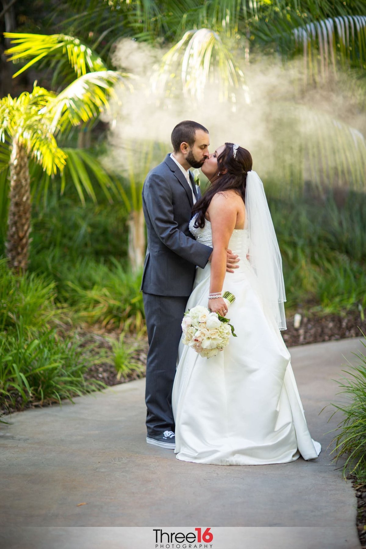 Bride and Groom stop along a path to share a kiss with beautiful foliage behind them at the Grand Tradition Estate wedding venue in Fallbrook