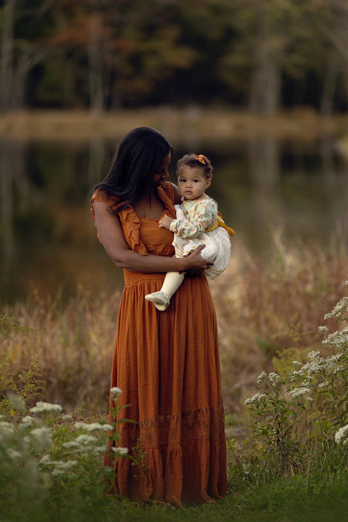 A mother in an orange dress stands by a lake holding her toddler daughter in a white dress on her hip New Jersey Family Photographer