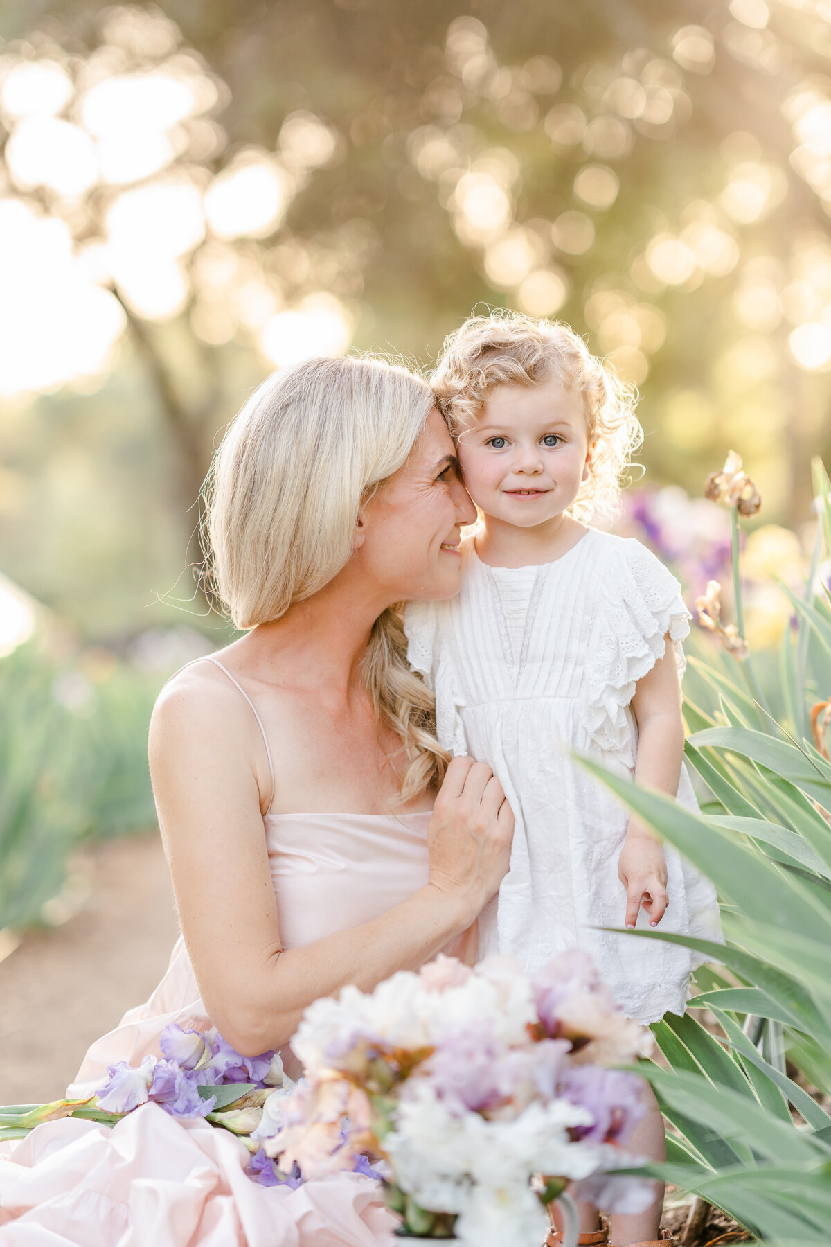 A mother sits while holding and placing her nose on her young daughter's check in a field of iris flowers photographed by Bay area photographer, Light Livin Photography.