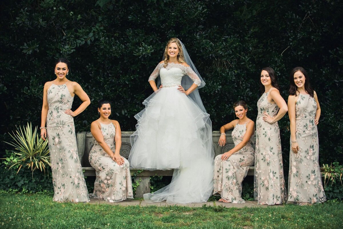 Bride in a white gown flanked by bridesmaids in patterned dresses, all smiling in a lush garden setting