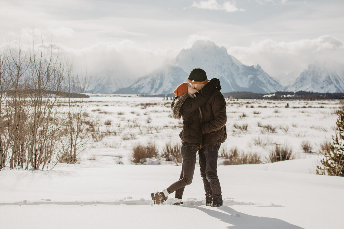 jackson hole photographers captures engaged couple kissing during their winter engagement session in Jackson Hole with the Grand Tetons behind them while it is snowing