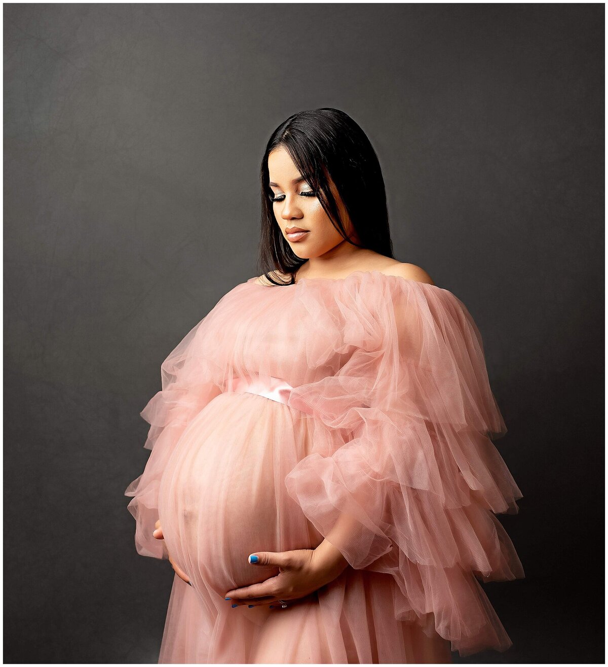 Pregnant woman wearing a beautiful  tulle dress poses with hand under belly and looking down slightly, showcasing her large belly.