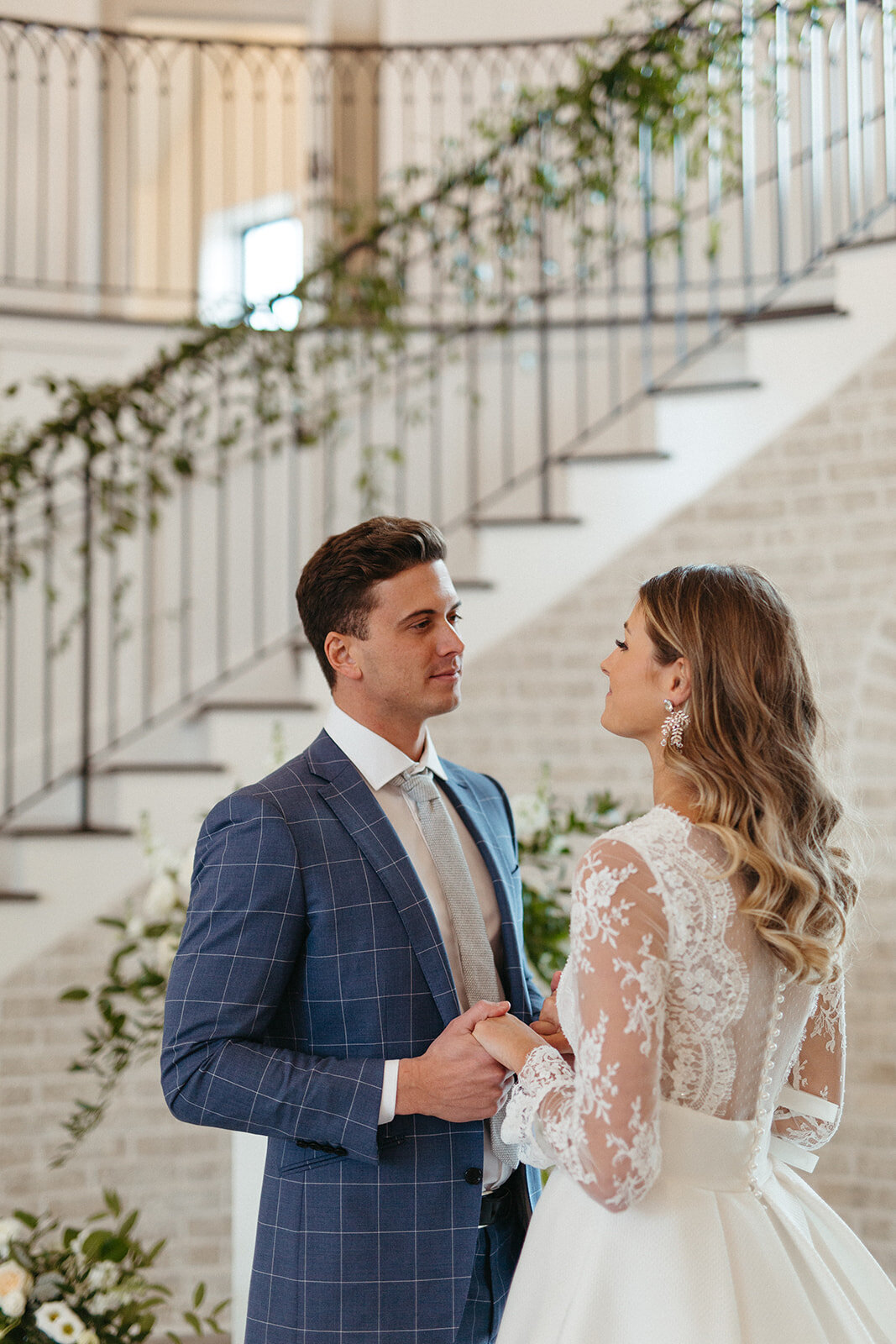Bride and groom in a blue suit and white wedding gown holding hands by a staircase wrapped in garland.